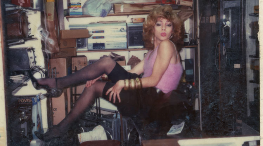 An old polaroid photograph of Venus Xtravagansa posing. She has a pink top, heels and gold bangles and is sitting in a crowded storage room.