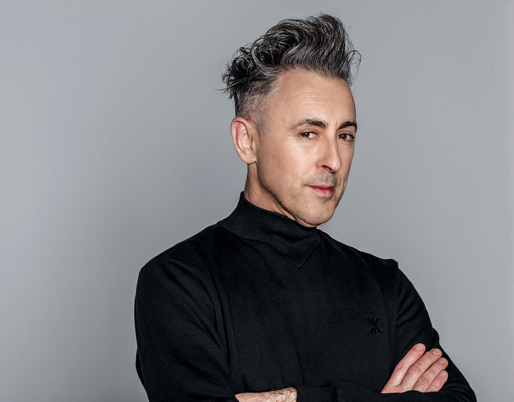 Actor Alan Cumming. He has his arms crossed, is looking directly into the camera and is wearing a black jumper.